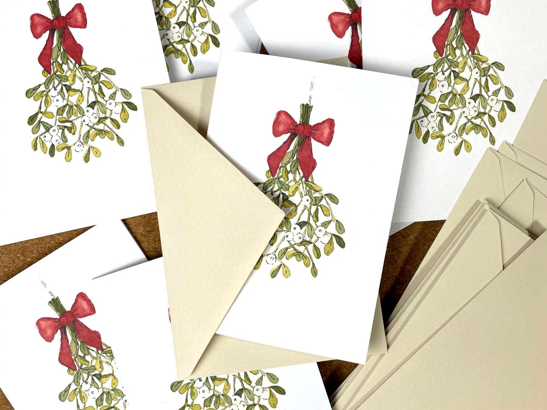 Mehrere Weihnachtskarten und beigefarbene Umschläge liegen auf einem Tisch. Die Karten sind mit einer Zeichnung eines Mistelzweigs mit einer roten Schleife bedruckt. Die Mistelzweig-Illustration wurde mit Aquarellfarben und Tusche erstellt.