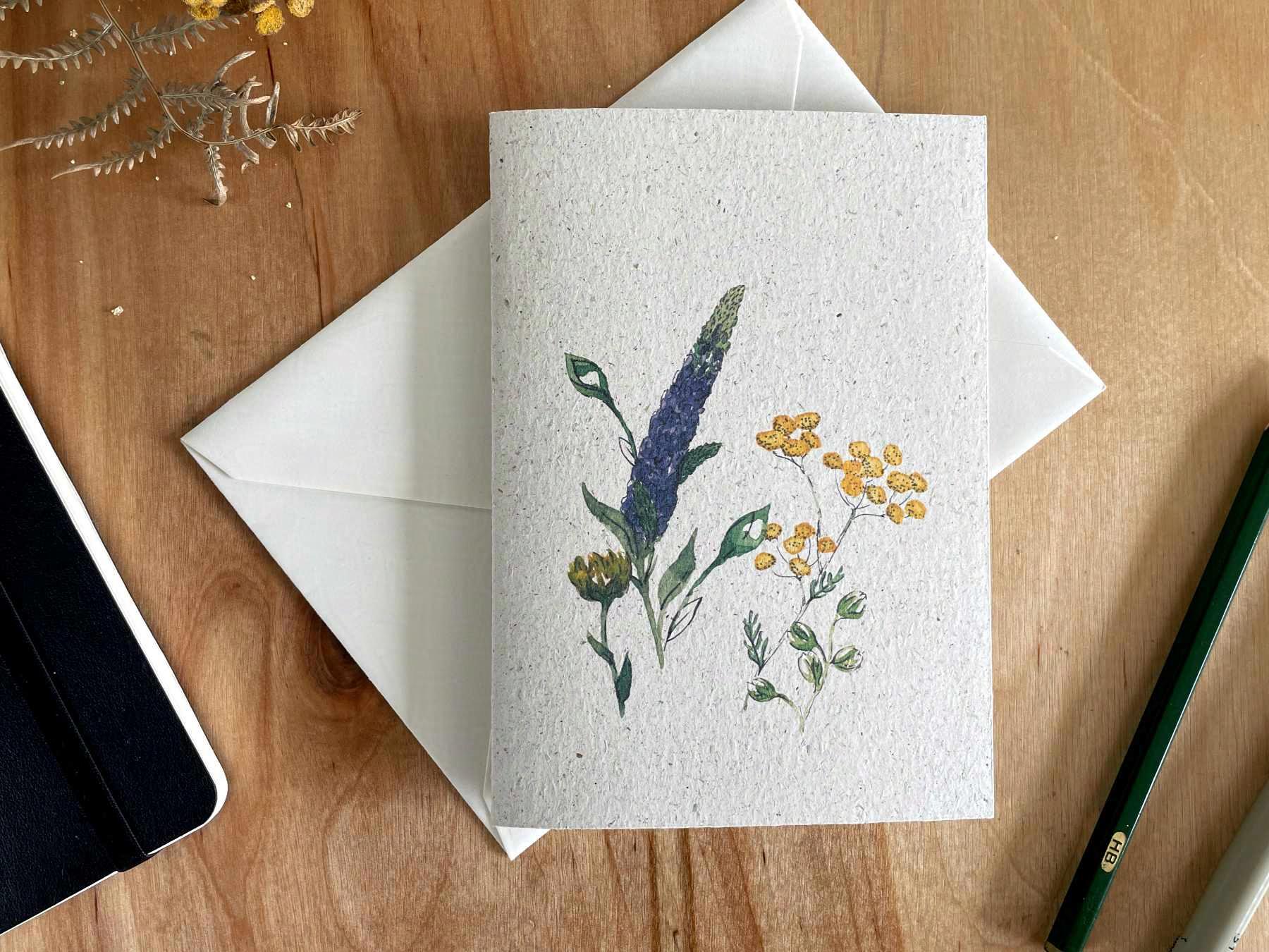 Eine Grußkarte aus Graspapier liegt mit einem weißen Umschlag auf einem Holztisch. Auf die Karte ist eine Zeichnung gedruckt, die mit Aquarellfarben und Tusche gemalte, lila und gelbe, sommerliche Wildblumen zeigt.