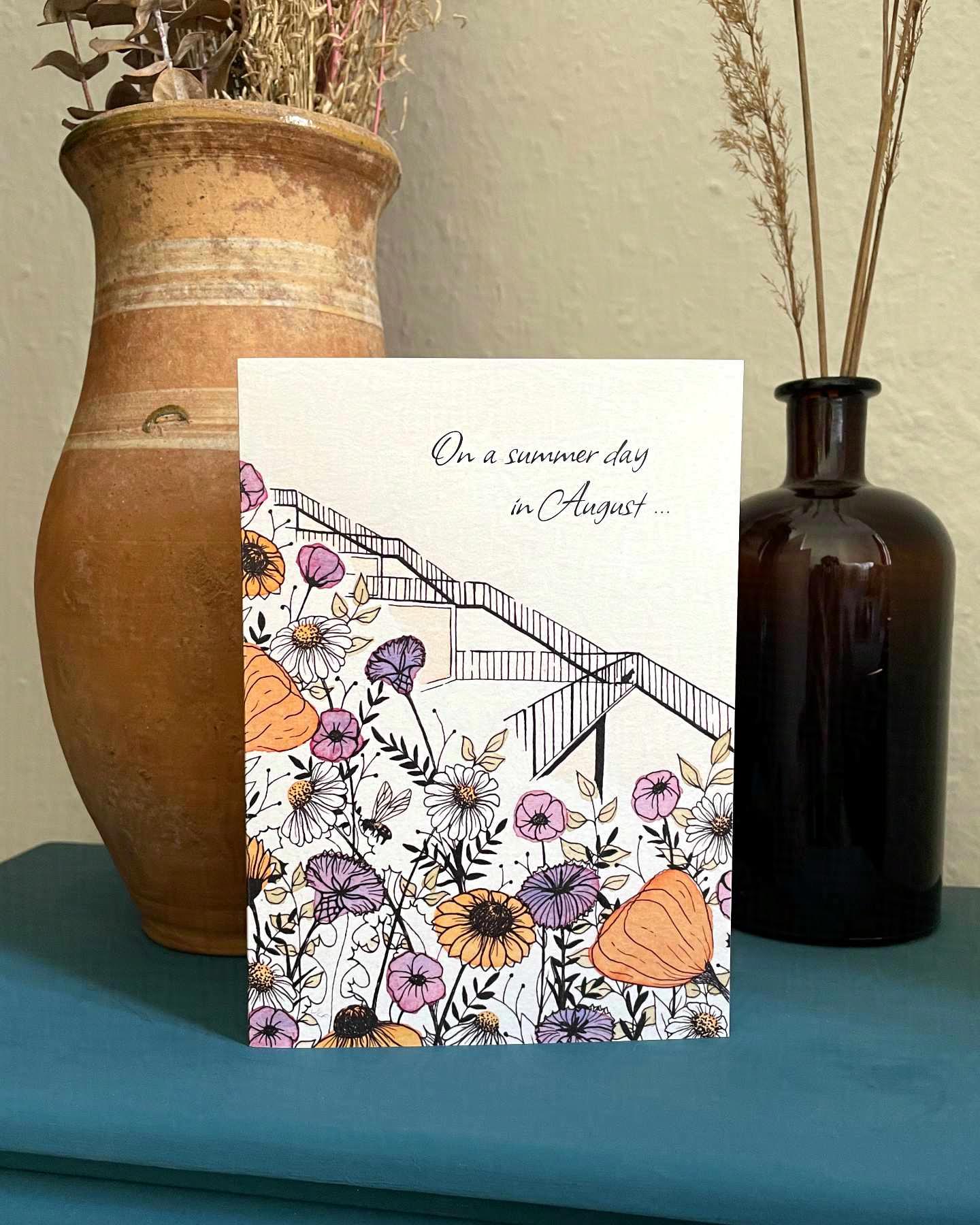 Eine mit Aquarellfarben und Tusche illustrierte Hochzeitseinladung steht vor zwei Blumenvasen auf einem kleinen petrolfarbenen Schrank. Auf der Karte sind Gräser und Wildblumen in Gelb-, Orange-, Rosa- und Lilatönen sowie eine Biene zu sehen.