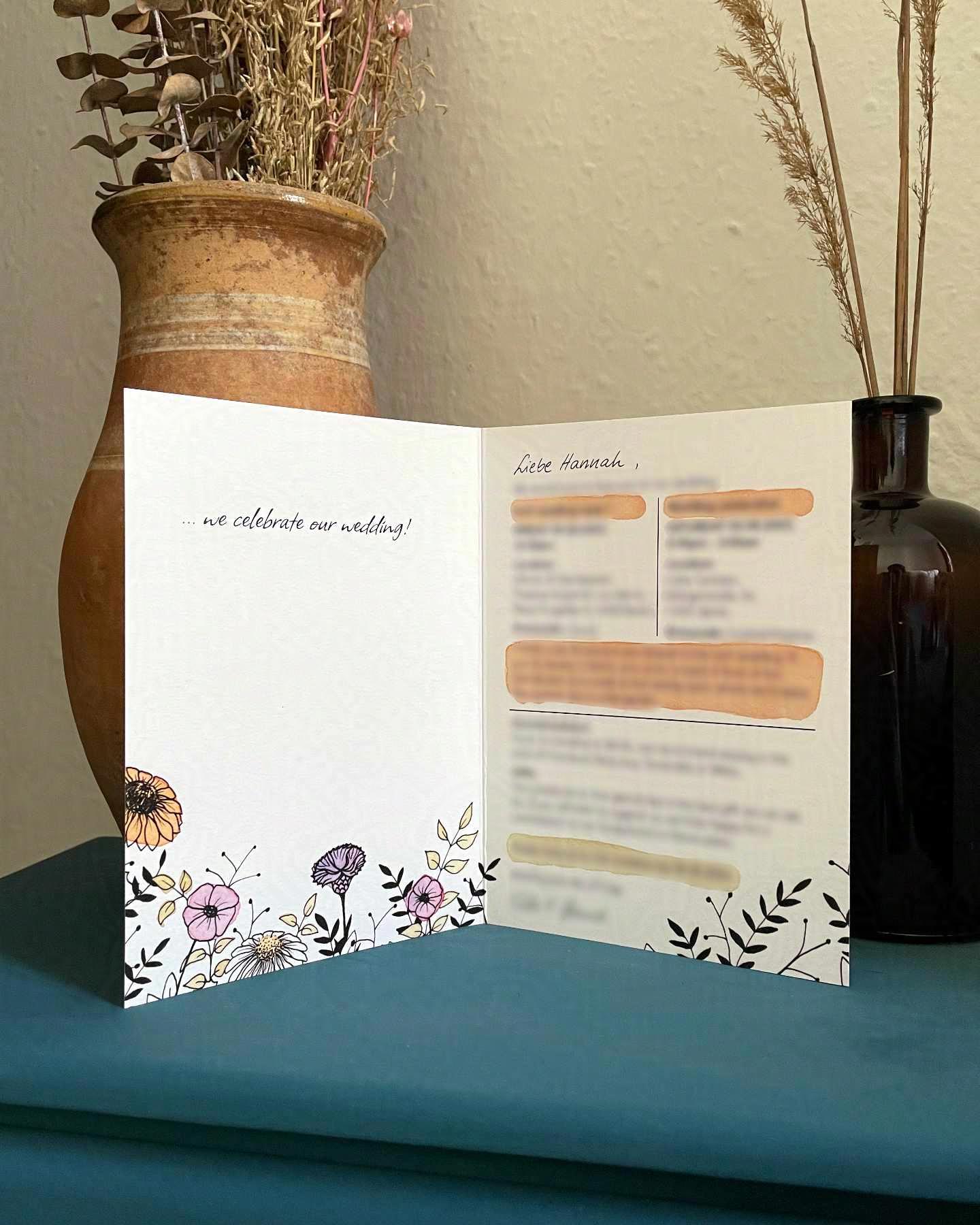 Eine von Hand illustrierte Hochzeitseinladung steht aufgeklappt vor zwei Blumenvasen auf einem kleinen petrolfarbenen Schrank. Auf den Innenseiten der Karte sind Gräser und Wildblumen zu sehen.