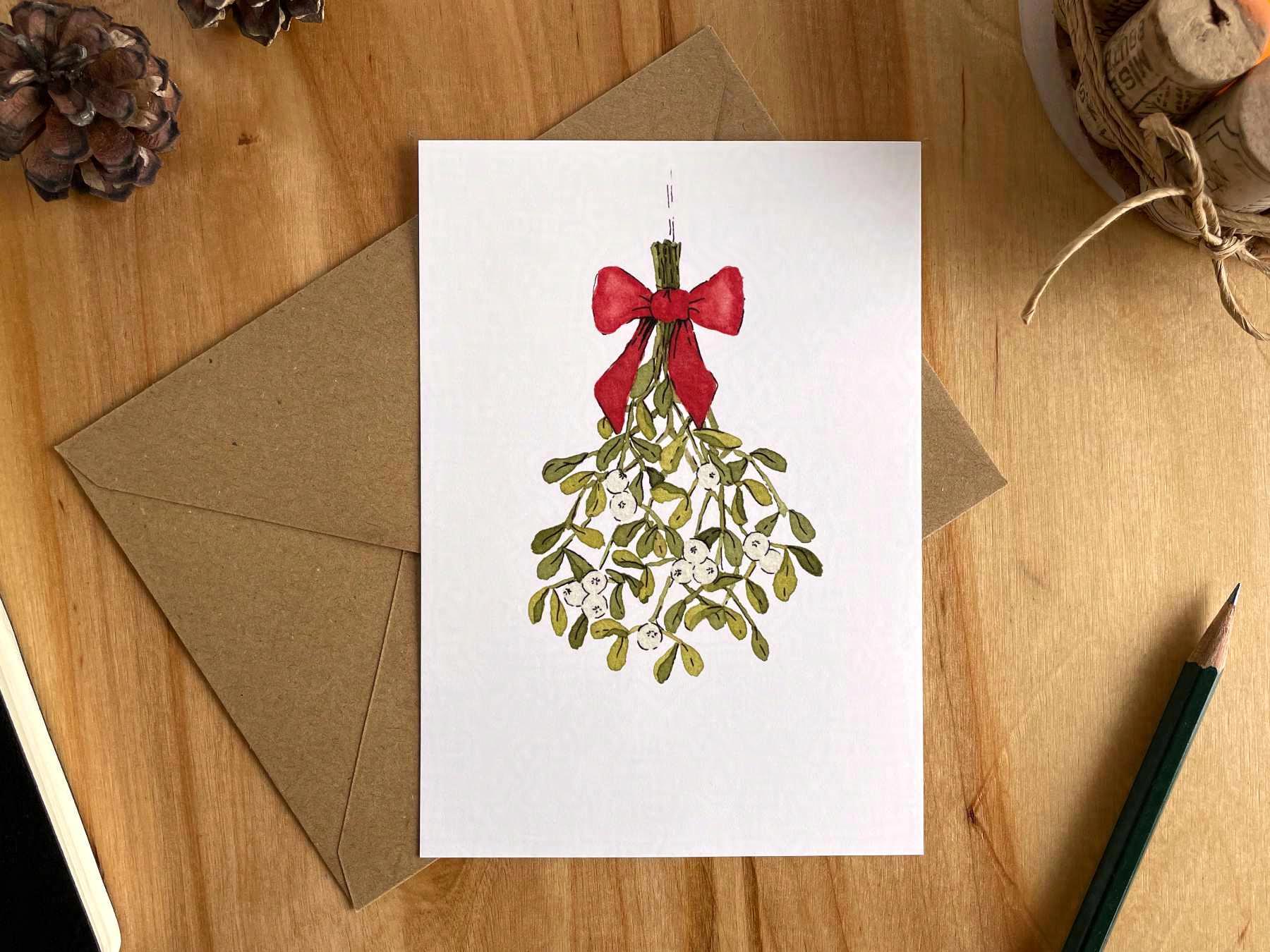 Eine Postkarte liegt mit einem braunen Umschlag auf einem Holztisch. Die Postkarte ist mit einer Zeichnung in Aquarellfarben und Tusche bedruckt, die einen Mistelzweig mit einer roten Schleife zeigt.