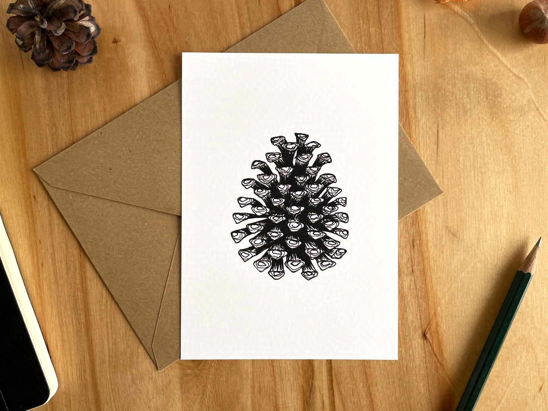 Eine Postkarte liegt mit einem braunen Umschlag auf einem Holztisch. Die Postkarte ist mit einer detaillierten Zeichnung eines Tannenzapfens bedruckt, der mit schwarzer Tusche gezeichnet wurde.