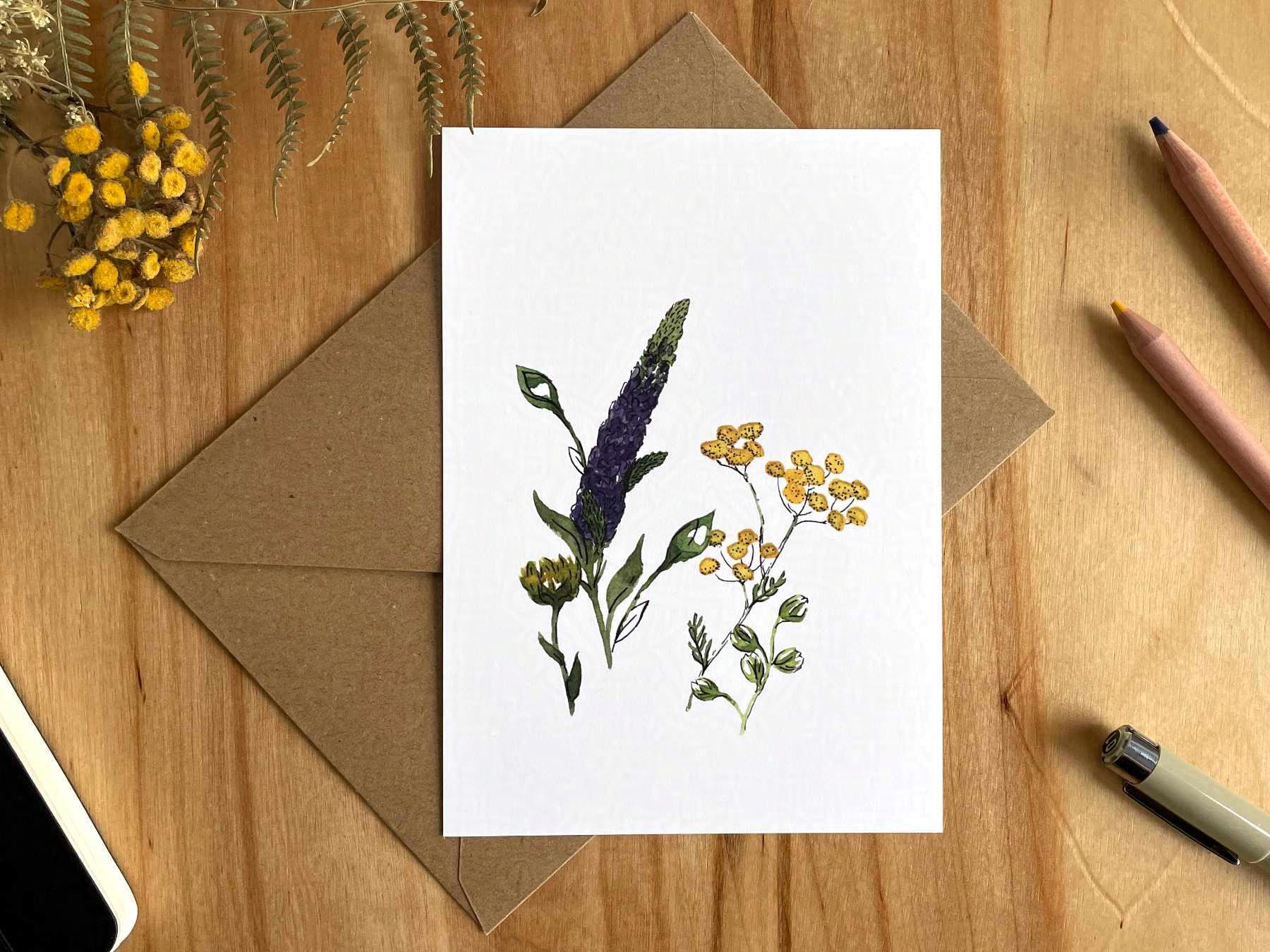 Eine Postkarte liegt mit einem braunen Umschlag auf einem Holztisch. Auf die Postkarte ist eine Zeichnung gedruckt, die mit Aquarellfarben und Tusche gemalte, lila und gelbe, sommerliche Wildblumen zeigt.