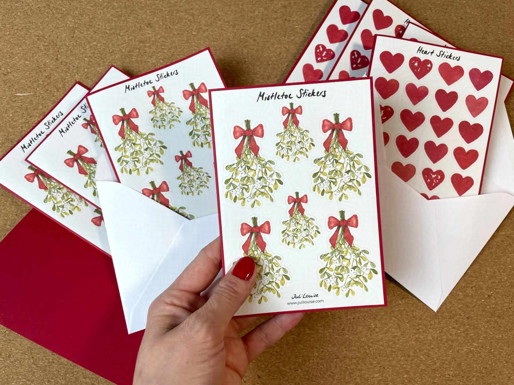 Mehrere Weihnachtskarten aus rotem Papier und weiße Umschläge liegen auf einem Tisch. Im Vordergrund wird eine der Karten in einer Hand gehalten. Auf der Karte sind Sticker angebracht, die mit Aquarellfarben und Tusche gezeichnete Mistelzweige zeigen.