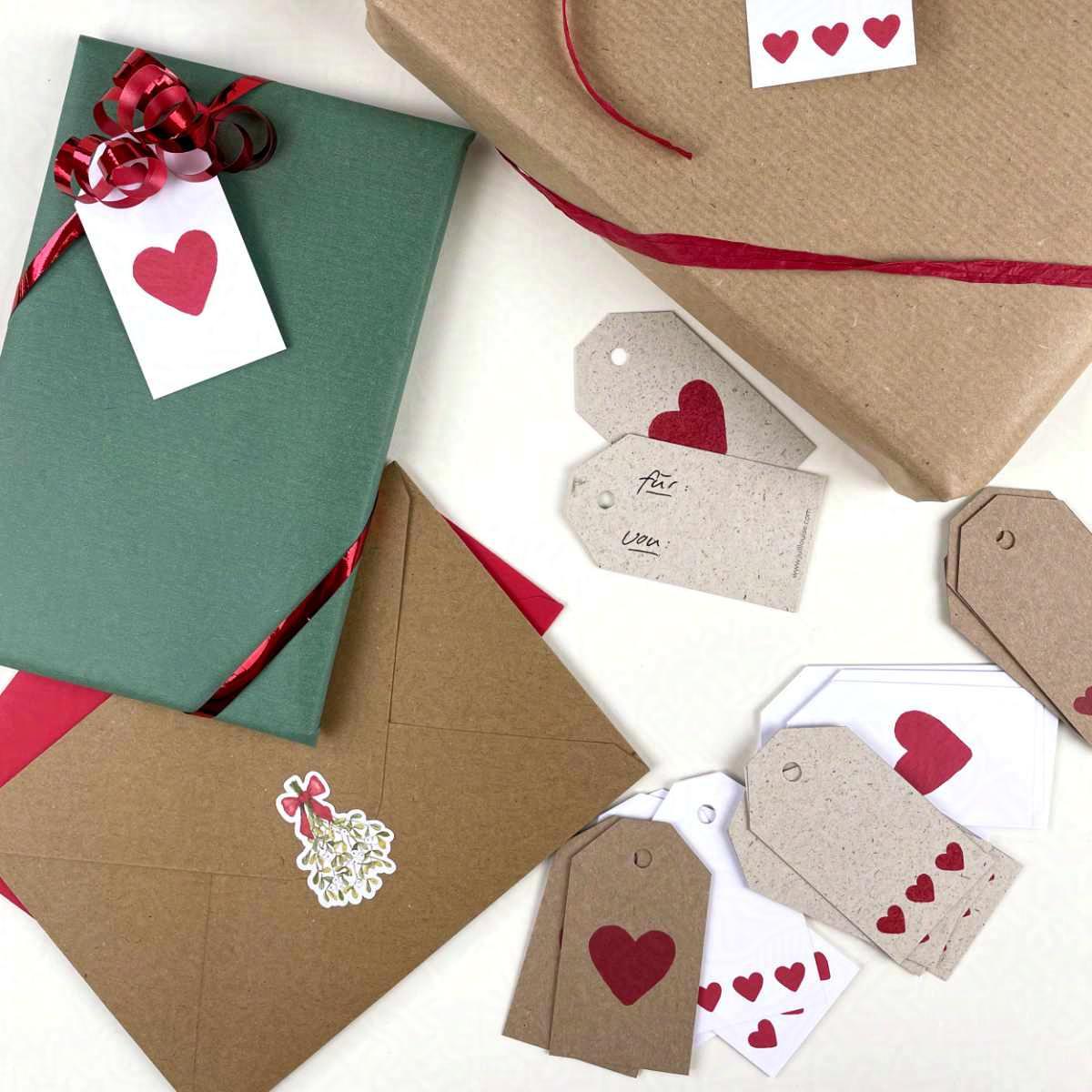 Geschenke, Briefe und Geschenkanhänger aus weißem Papier, Graspapier und Kraftpapier liegen auf einem Tisch. Die Anhänger sind mit roten Herzen in verschiedenen Größen bedruckt.