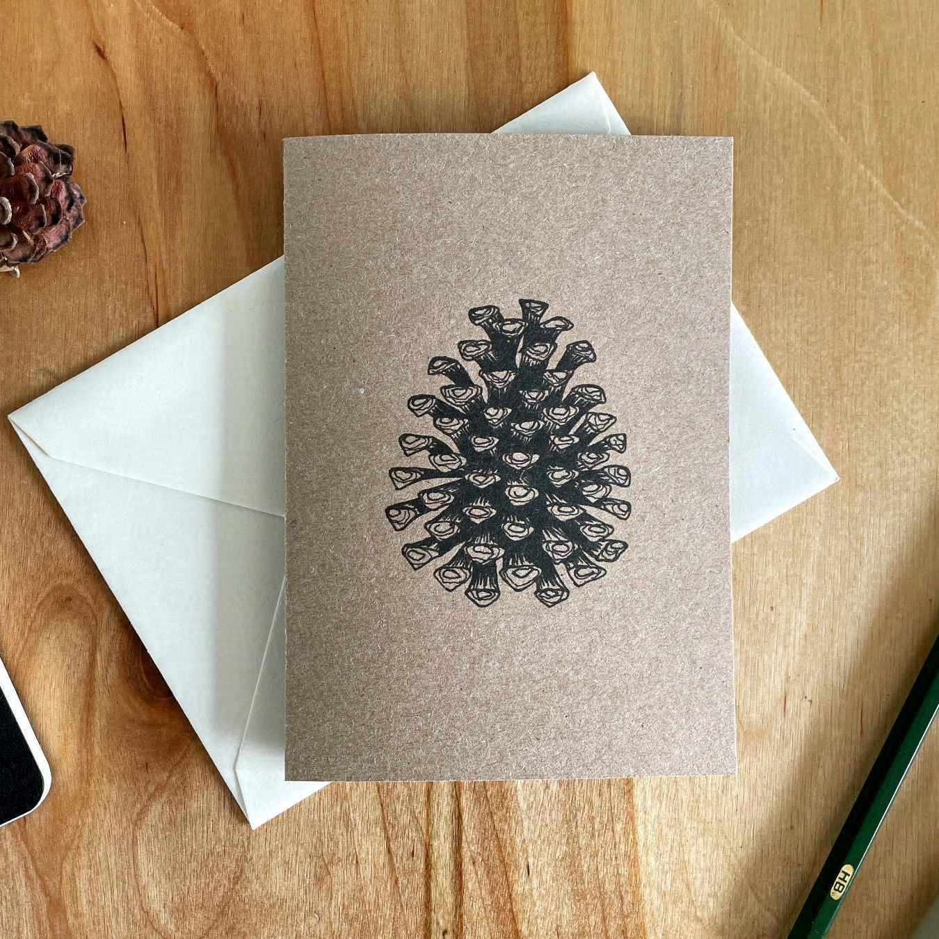 Eine Grußkarte aus Kraftpapier liegt mit einem weißen Umschlag auf einem Holztisch. Die Karte ist mit einer Zeichnung von einem Tannenzapfen bedruckt, der mit schwarzer Tusche gezeichnet wurde.