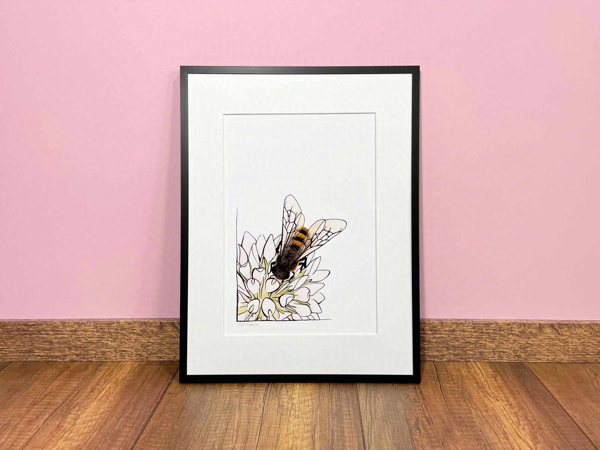 Ein schwarzer Bilderrahmen steht auf einem Holzfußboden an eine hellrosa Wand gelehnt. In dem Rahmen befindet sich ein Druck einer Aquarell- und Tusche-Zeichnung, die eine Biene auf einer Blüte zeigt.