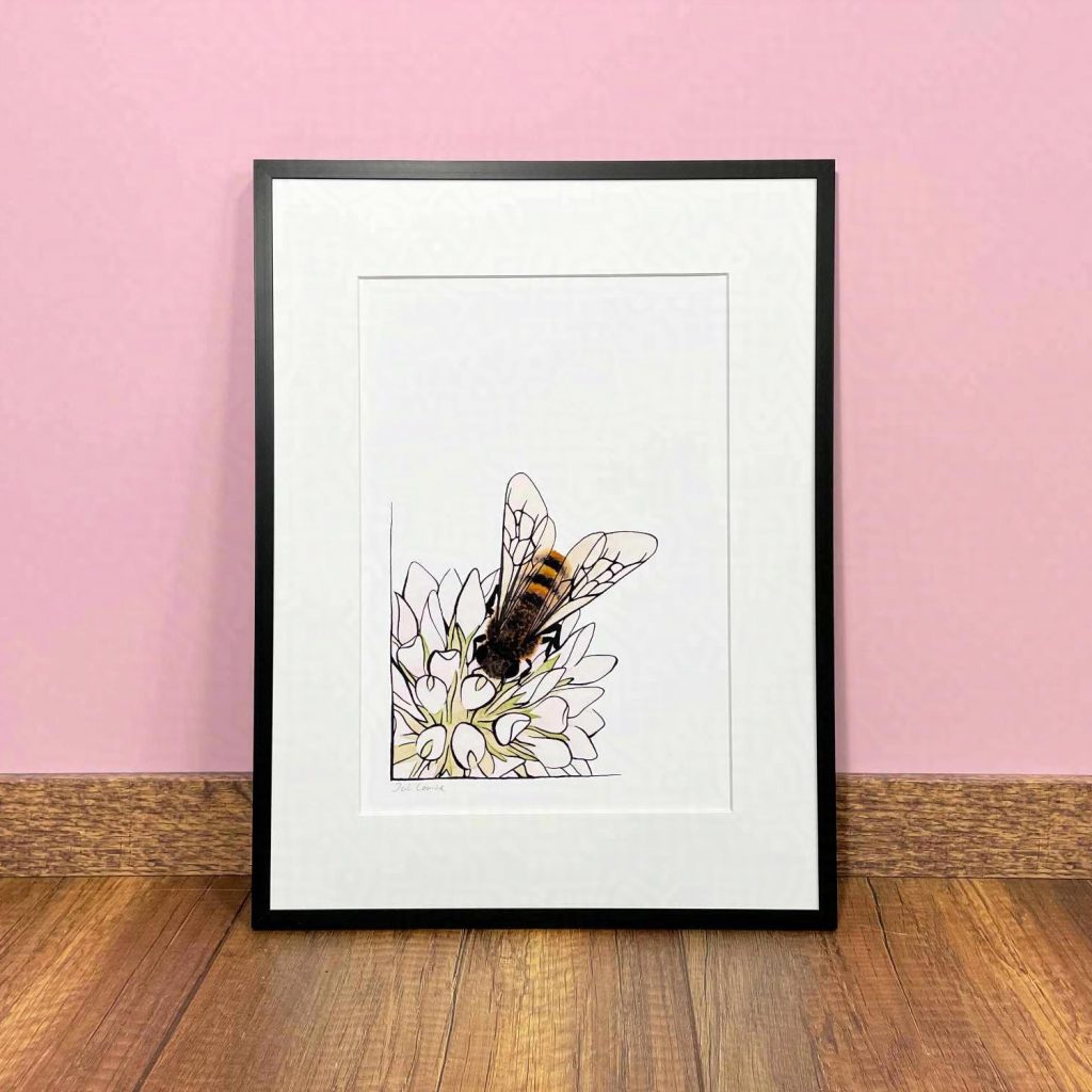 Ein schwarzer Bilderrahmen steht auf einem Holzfußboden an eine hellrosa Wand gelehnt. In dem Rahmen befindet sich ein Druck einer Aquarell- und Tusche-Zeichnung, die eine Biene auf einer Blüte zeigt.