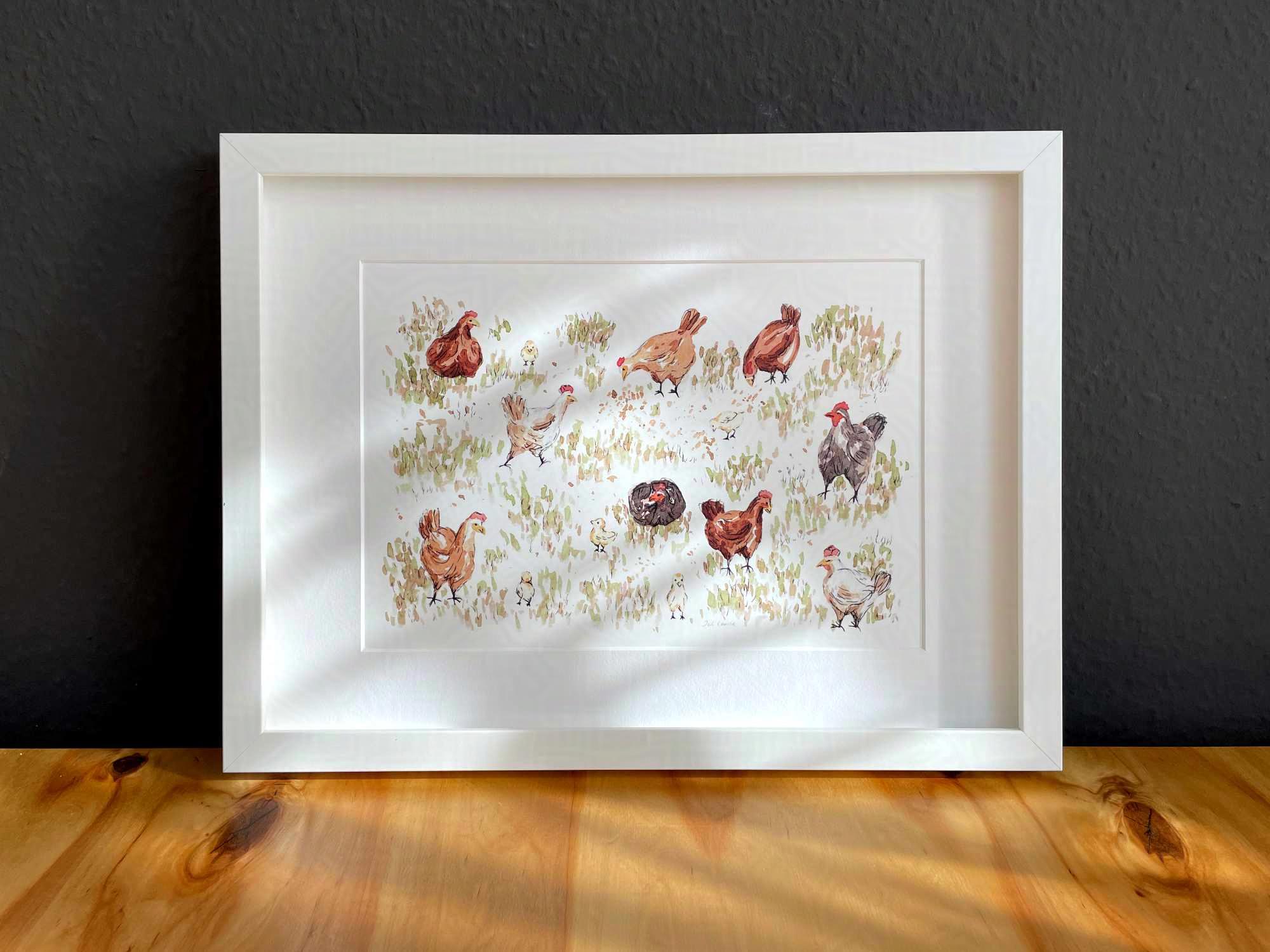 Ein weißer Bilderrahmen mit einem Druck darin steht auf einem Holztisch an eine dunkle Wand gelehnt. Der Druck zeigt eine Aquarell- und Tusche-Illustration von freilaufenden Hühnern.