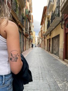 Im Vordergrund ist die Rückseite des angewinkelten Arms einer jungen Frau zu sehen, auf dem sie ein Tattoo trägt. Das Tattoo zeigt eine mit feinen, schwarzen Linien erstellte Zeichnung von verschiedenen Wildblumen, Gräsern und einer Biene.