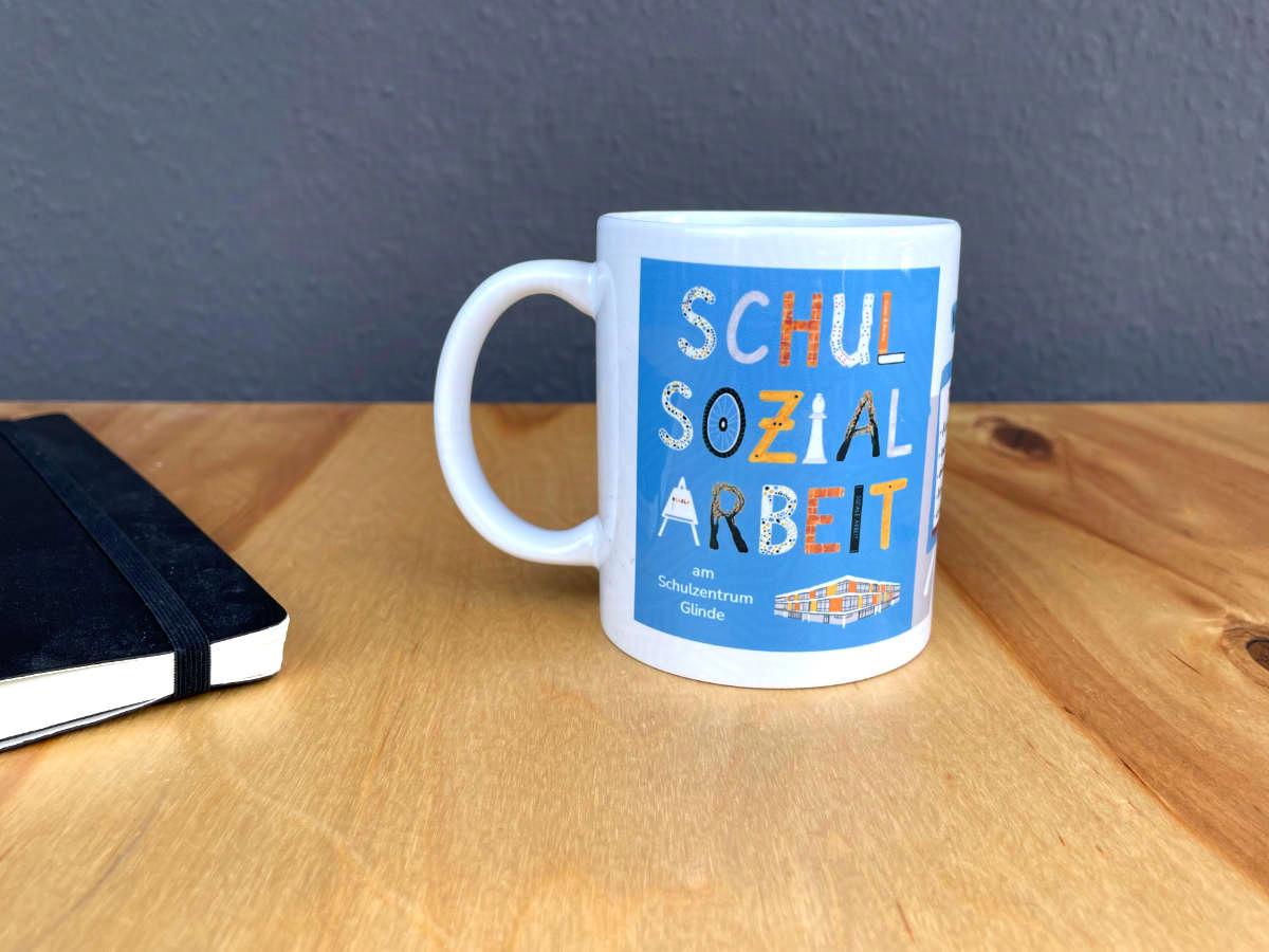 Eine Tasse steht auf einem Holztisch. Die Tasse ist mit einer Illustration für die Schulsozialarbeit am Schulzentrum Glinde bedruckt.