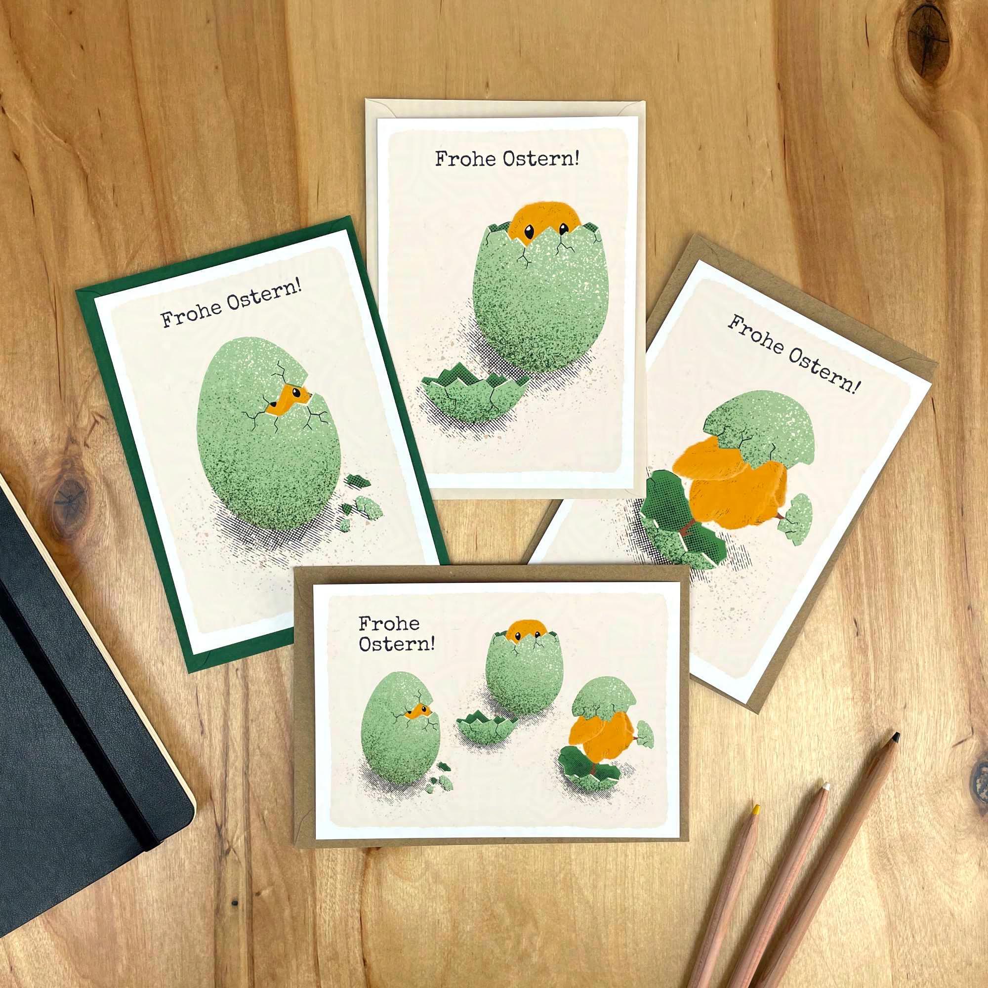 Auf einem Holztisch liegen vier Postkarten, die Illustrationen von Osterküken zeigen, die gerade aus ihren Eiern schlüpfen. Unter den Karten liegen außerdem jeweils farblich passende Umschläge.