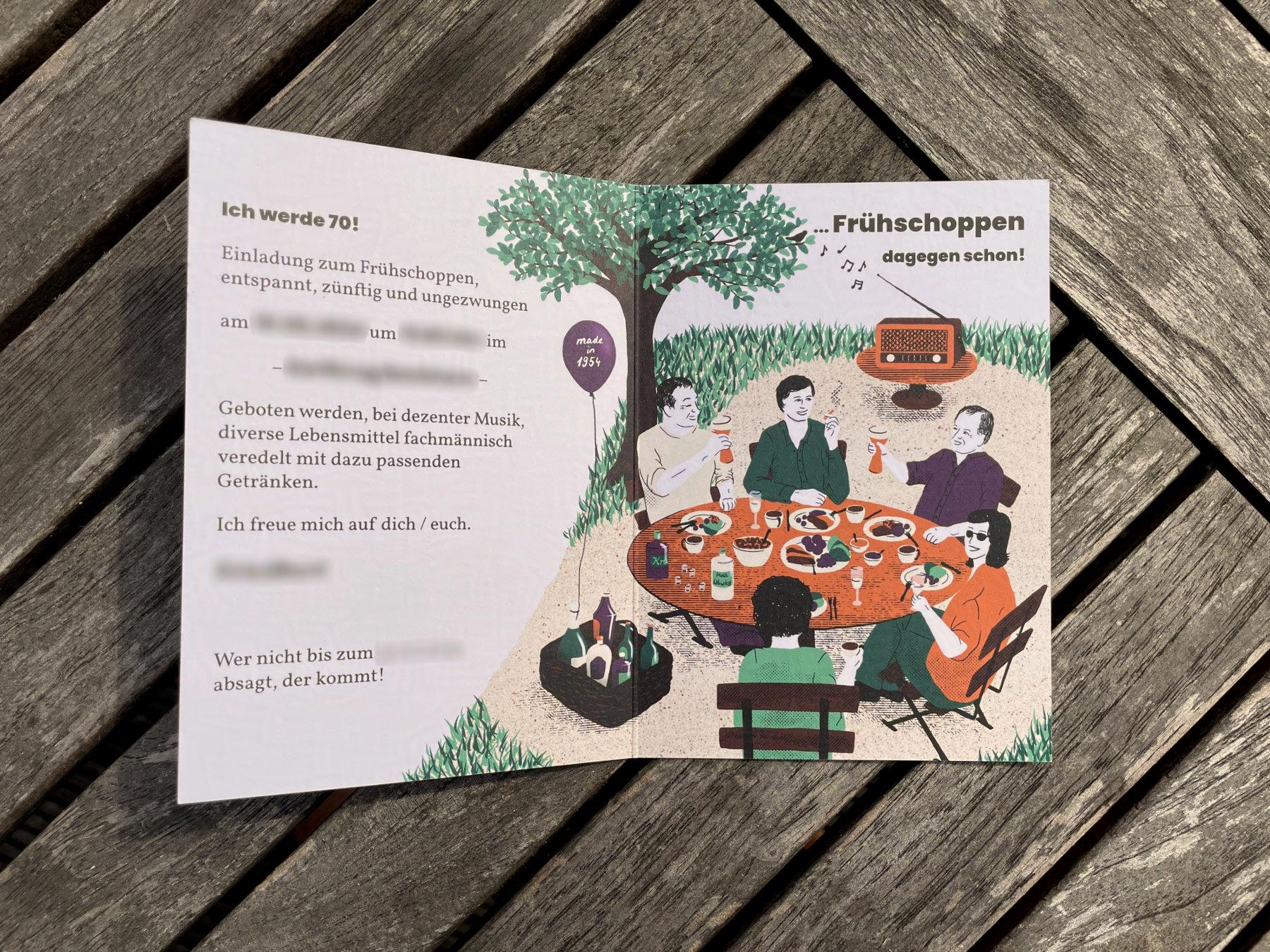 Eine illustrierte, aufgeklappte Geburtstagseinladung liegt auf einem Tisch. Die Karte zeigt eine Gruppe Menschen, die gemeinsam an einem Gartentisch sitzen und essen.