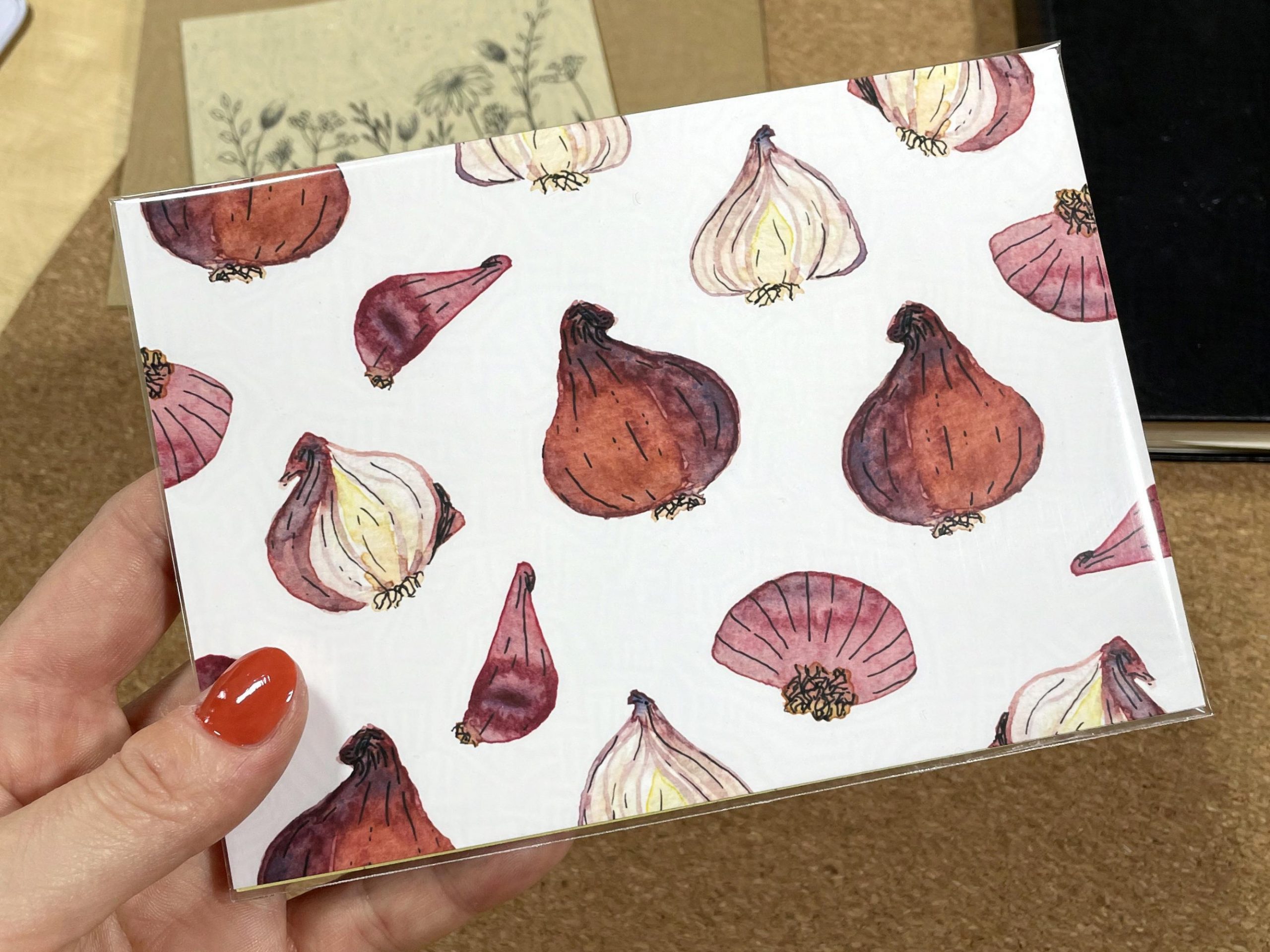 Ein kleiner Druck, auf dem rote Zwiebeln abgebildet sind, wird in einer Hand gehalten. Die Zwiebeln wurden mit Aquarellfarben und Tusche illustriert.